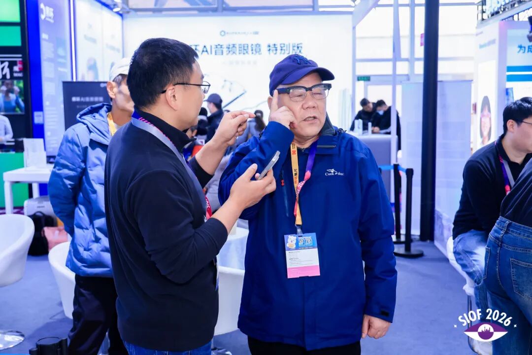 At an AI smart glasses event, two men converse amidst a bustling exhibition room filled with technology displays.